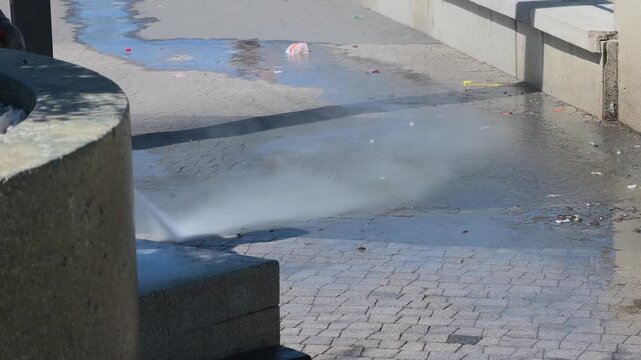 Unrecognizable industrial groundskeeper wearing workwear deeply cleaning urban pedestrian plaza using heavy duty power washer machinery with intense water jet spray under bright sunlight wide shot
