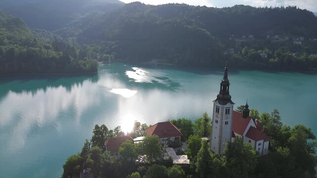 Aerial of the Assumption of Mary Church tower on Bled Island, Slovenia. Beautiful cinematic drone footage of the historic church with the sun reflecting on the turquoise lake water in the background.