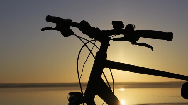 A dark silhouette of bicycle handlebars stands against a bright golden sunset reflecting over calm water. Concept of peaceful outdoor recreation, active lifestyle, and summer travel.