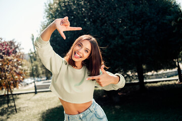 Naklejka premium Playful young woman in crop top and jeans framing a selfie with her hands in a sunny park, smiling and sticking out her tongue in a carefree outdoor lifestyle portrait