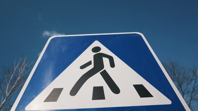 A blue and white pedestrian crossing sign stands clearly against a bright, sunny blue sky. Bare winter tree branches frame the edges of the traffic safety marker during the day.