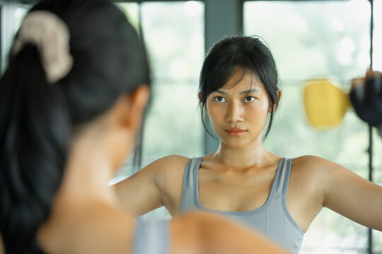 Asian woman in gray sportswear holds a yellow dumbbell at her shoulder and studies her reflection in a gym mirror.