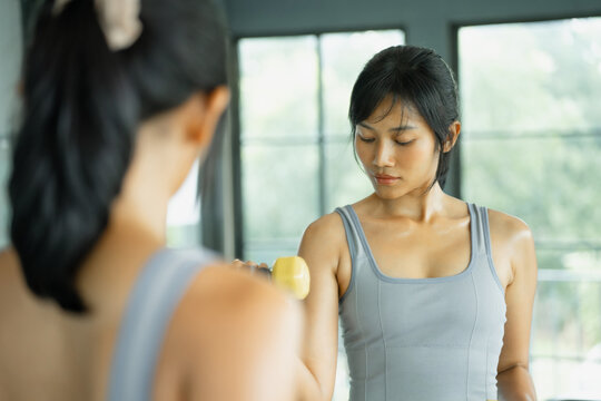 Asian woman in gray sportswear trains on a cardio machine in side profile by large sunlit gym windows with greenery outside.