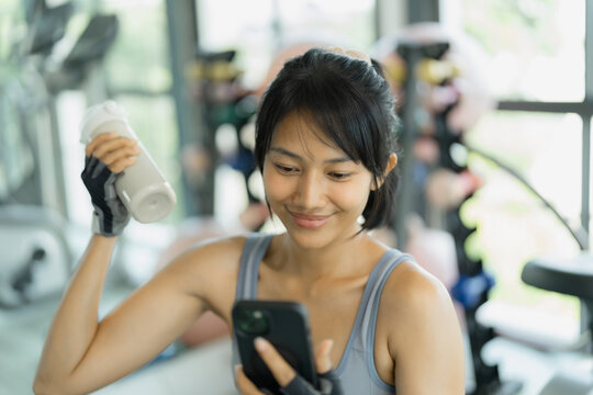 Asian woman in gray sportswear smiles at her phone while holding a shaker bottle of protein drink during a gym break.
