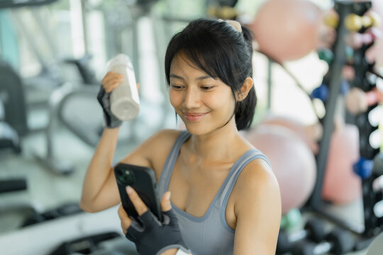 Asian woman in gray sportswear does a dumbbell curl with a yellow dumbbell, focused and looking down, with exercise balls behind her.