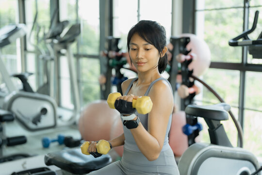 Asian woman in gray sportswear curls yellow dumbbells during a strength workout with gym equipment behind her.