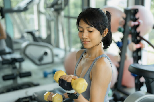 Asian woman in gray sportswear does a dumbbell curl with a yellow dumbbell, looking down with focus among exercise balls.