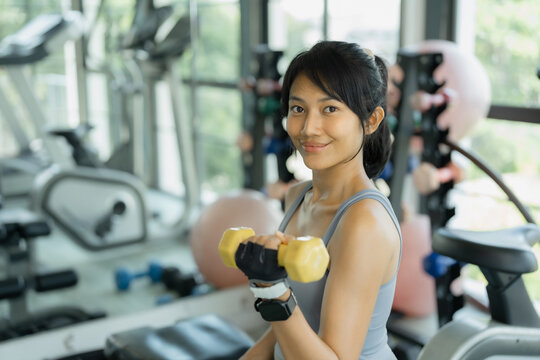 Asian woman in gray sportswear smiles at the camera while holding a yellow dumbbell during a workout in a bright gym.