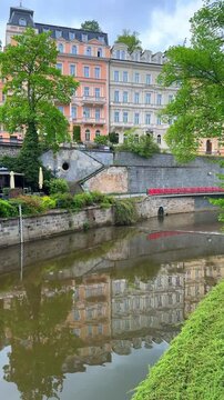 The Tepla river in Karlovy Vary. The buildings stand tall with reflections on the water. Nature surrounds the area, creating a lovely place to visit.