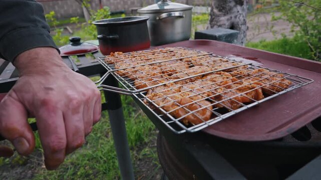 A person holds a metal grill grate loaded with marinated meat. Cooking pots rest on an outdoor table in the background. The scene depicts preparations for a backyard barbecue.