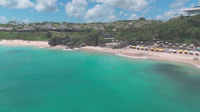 A smooth aerial drone shot captures Dreamland Bay, Bali from farther out over the ocean, revealing a broader view of the coastline and beach below. The expansive turquoise waters fill the foreground,
