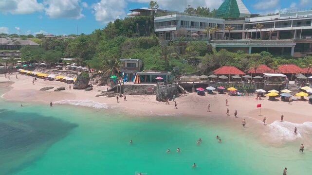 A cinematic drone shot glides just offshore in Dreamland Bay, Bali, looking back toward the beach where rows of colourful umbrellas dot the golden sand.
