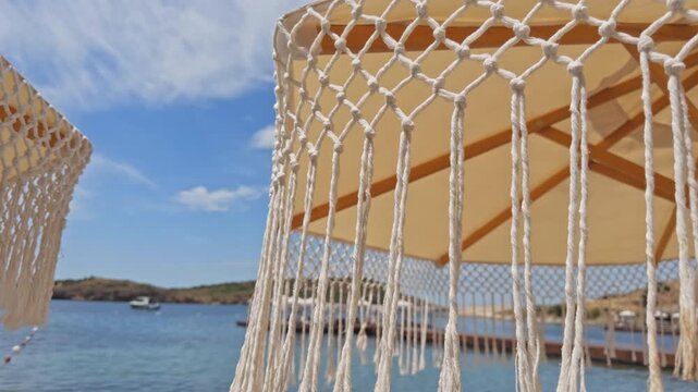 High-quality 4K close-up shot of a white handmade macrame sun umbrella at a luxury beach resort. Features empty sunbeds, sandy beach, and a beautiful summer resort background.