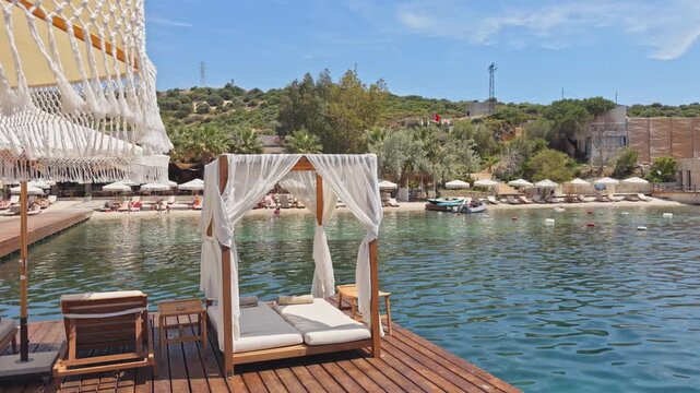 Slow pull-back wide shot of white canopy daybed on wooden pier over turquoise Aegean bay, luxury beach club with sun loungers in background,