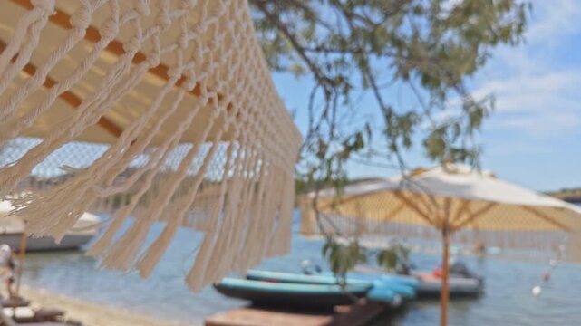 Close-up shot with shallow depth of field showing macrame fringes of a boho beach umbrella waving in the wind at a luxury summer resort with boats in the blurred background.
