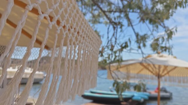 Close-up shot with shallow depth of field showing macrame fringes of a boho beach umbrella waving in the wind at a luxury summer resort with boats in the blurred background.