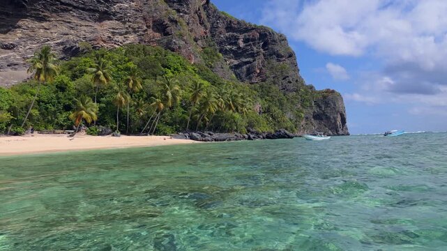 Playa Fronton secluded beach with turquoise water, palm-lined cove beneath dramatic rocky cliffs and boats anchored offshore under partly cloudy skies. Dominican Republic. Aerial drone low view