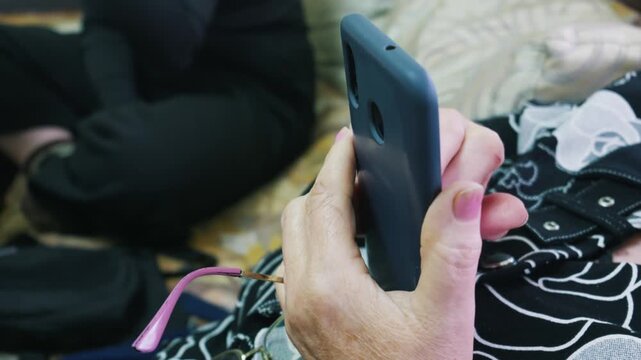 Senior woman holds a blue smartphone in a protective case with pink glasses tucked between her fingers. She wears a patterned shirt and pink nail polish. Senior technology usage concept.