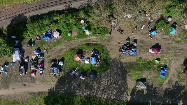 Overhead view of a sprawling homeless encampment with scattered tents, blue tarps and debris along worn dirt paths beside active railroad tracks in Roseburg, Oregon, USA. Aerial drone top down.