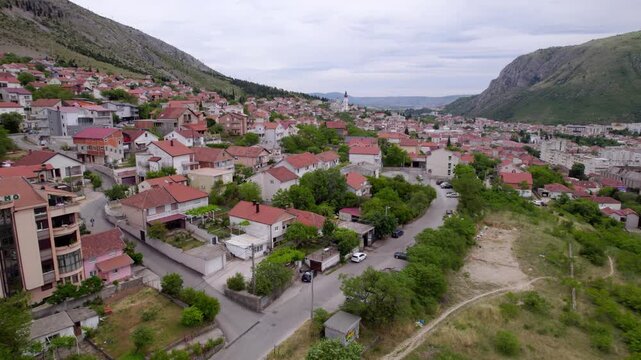Aerial establishing across Mostar neighborhoods between mountain slopes with winding local roads, orange subdued rooftops, Bosnia and Herzegovina