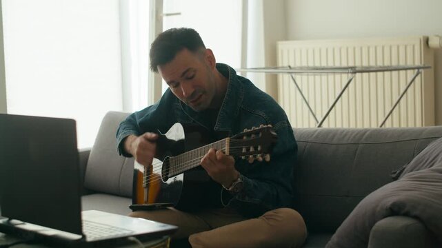 An authentic young man practices acoustic guitar at home using a laptop for remote learning. Perfect for wellness, hobby, and digital nomad lifestyle content.