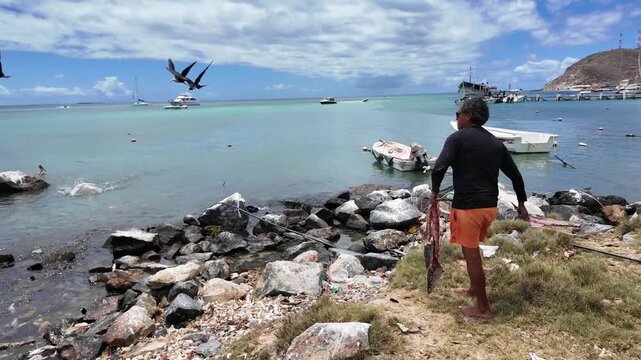 Ecological cycle showing a local worker sharing fresh fish remnants with frigatebirds and gulls on a tropical shore.