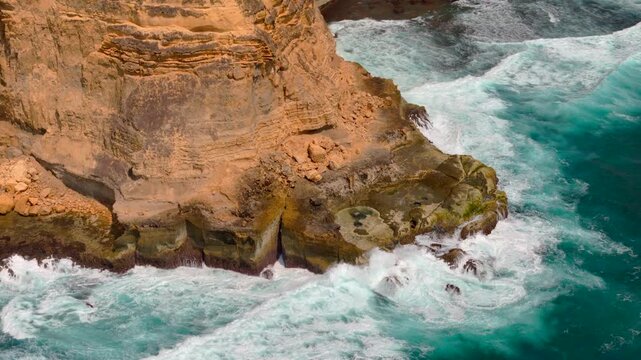 Drone view tracks a golden rocky cliff face as turquoise waves crash into the coast creating white foam for dramatic geology ocean power and travel landscape themes