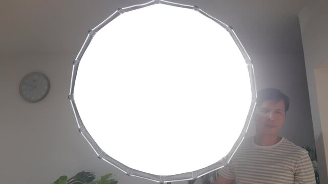 Over-the-shoulder shot of a content creator turning on a large round studio softbox light, illuminating the room for video production.