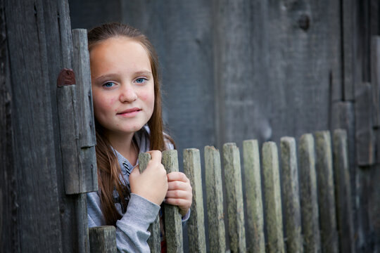 Young girl looking through a wooden fence beside a rustic building, holding the gate and smiling softly at the camera.