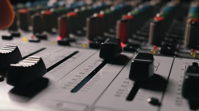 Hand of an audio engineer slides a fader up on a professional soundboard in a recording studio. Red and yellow lights illuminate the console buttons. Sound engineering concept included.
