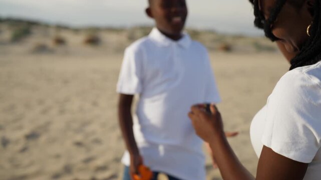 Cheerful african american boy learning to juggle oranges with his mother on a sandy beach, enjoying a sunny day outdoors and sharing a happy, playful moment together