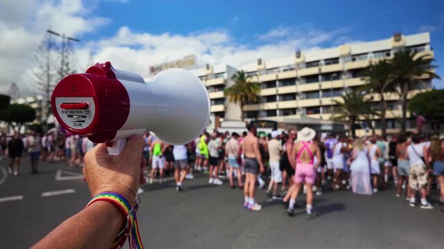 Point of view of a person holding a megaphone with a rainbow wristband at a gay pride street parade, surrounded by a large crowd celebrating LGBT rights and freedom during pride month