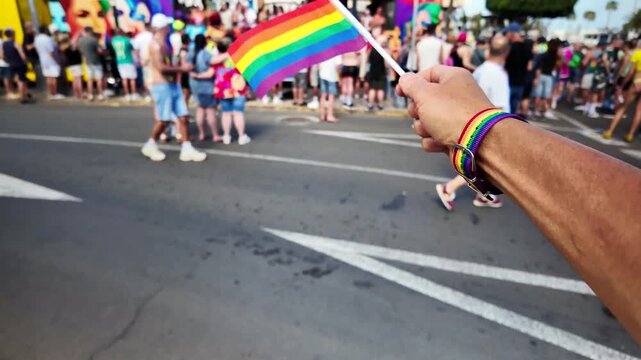Point of view shot of a person's hand holding a waving rainbow flag with a pride bracelet, marching during a gay pride parade on a sunny day with a crowd in the background