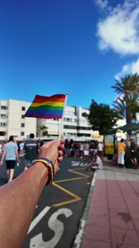 Point of view of a person holding a small waving LGBTQ+ rainbow flag during a sunny gay pride parade on a city street, showing support for the community and celebrating diversity and equality