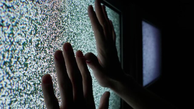 Human hands are gently pressed against the illuminated screen of a vintage cathode-ray tube television. The display projects a mesmerizing pattern of white noise and analog static, creating