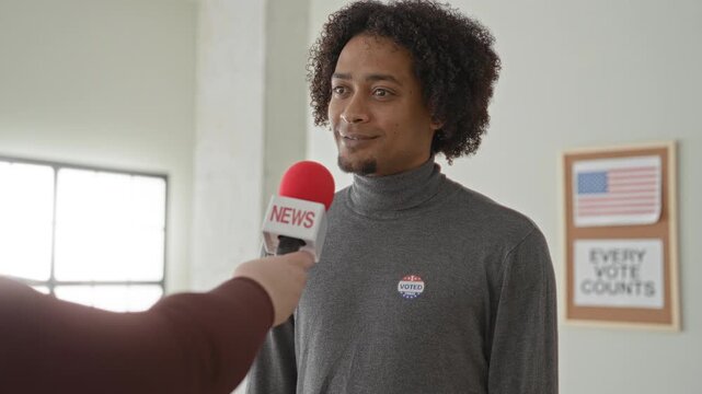 Man speaks into a red news microphone at a polling station, wearing an i voted sticker and answering questions about the elections; civic pride.