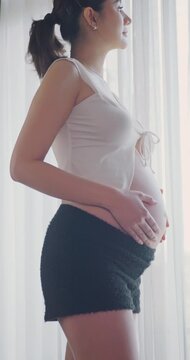 Close up of a pregnant woman standing by a window and touching her belly.