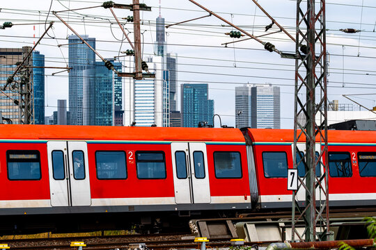 Red train passes across Frankfurt railway tracks, with Germany skyline towers, station wires and urban infrastructure layered behind the carriage windows