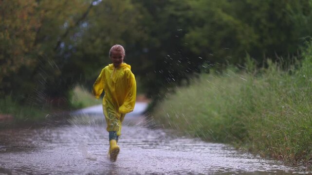 Happy An eight-year-old girl wearing yellow rubber boots and a yellow raincoat Jumping in a puddle after a summer rain.  feral kid summer,