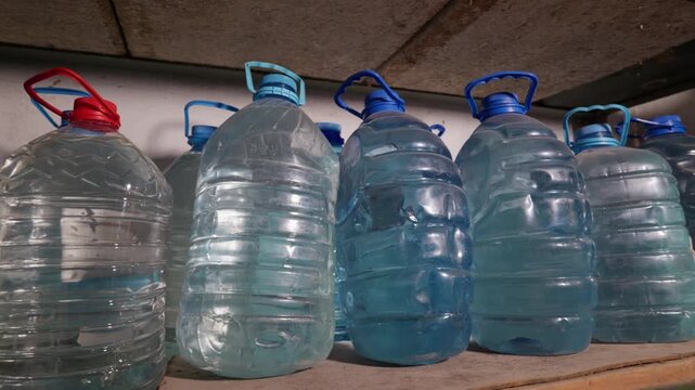 Rows of large plastic water jugs stand on a dusty concrete shelf in a dark storage room. Several blue and red capped containers are filled with fresh drinking water for emergency supply.