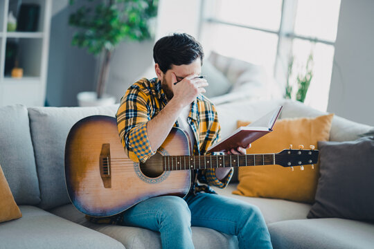 Frustrated songwriter with acoustic guitar reading notebook on sofa in living room, rubbing his forehead while struggling to compose lyrics and find inspiration at home