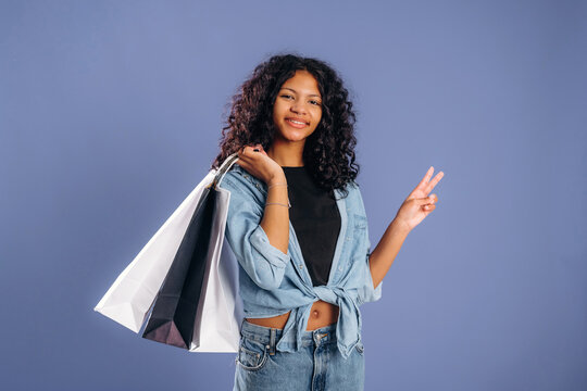 Young African American woman with curly hair holds shopping bags and makes a peace sign against a solid blue background in a casual outfit