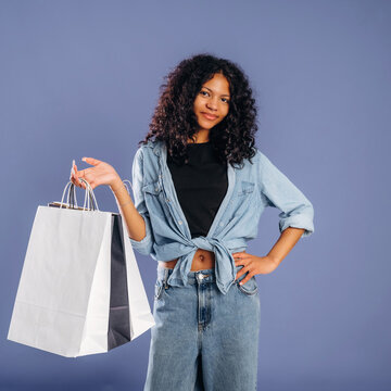 Young woman with curly hair wearing denim shirt and jeans holds shopping bag against a solid color background, showcasing a casual fashion style and shopping experience
