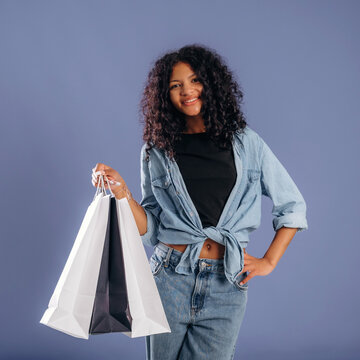 Young African American woman with curly hair smiles while holding shopping bags in front of a solid color background, wearing a casual outfit with rolled-up sleeves