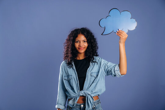 Young African woman with curly hair holds a blue speech bubble cutout against a solid purple background, wearing a denim shirt and casual attire