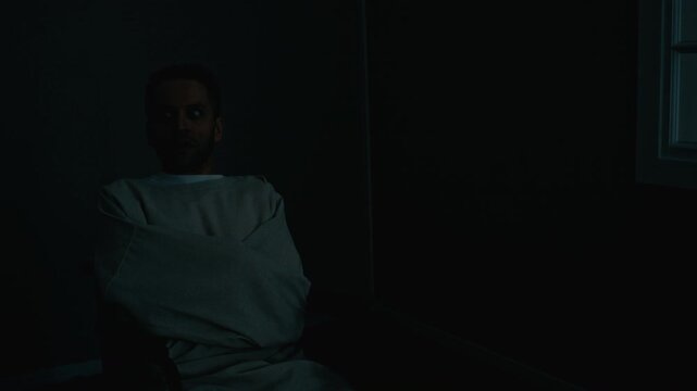 Medium shot of middle aged Caucasian man wearing straitjacket sitting in wheelchair, looking around anxiously in dark room with flickering light. Perfect for horror film or mental health documentary