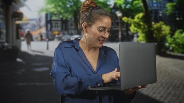 Woman brunette holding laptop and typing with fingers on keyboard on cobblestone street near trees and buildings; concentration productivity.