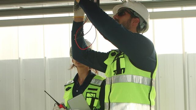 Engineers with safety vest and helmet work with electrical instrument on photovoltaic solar panels. Technicians discuss together. Woman use walkie talkie while man use multimeters to check solar power