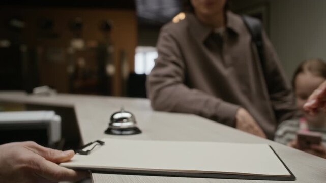 Cropped shot of woman checking in at hotel reception desk with family, handing passport and signing registration form. Ideal for travel agency promos and hospitality marketing