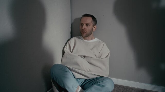 Dolly in of Caucasian man wearing straitjacket sitting in corner of psychiatric ward looking paranoid as shadows loom. Clip is perfect for mental health awareness and medical documentaries